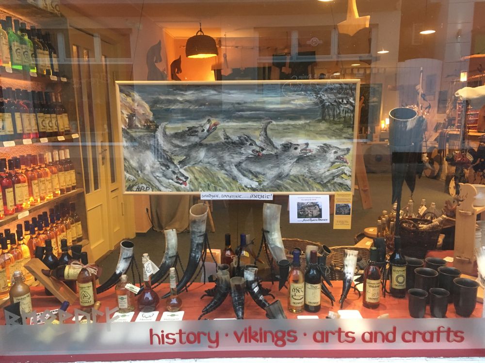 In einem Schaufenster hängt über einer Warenpräsentation von Trinkhörnern und Met ein Bild, das gemalt ein wildes Rudel Wölfe zeigt.
