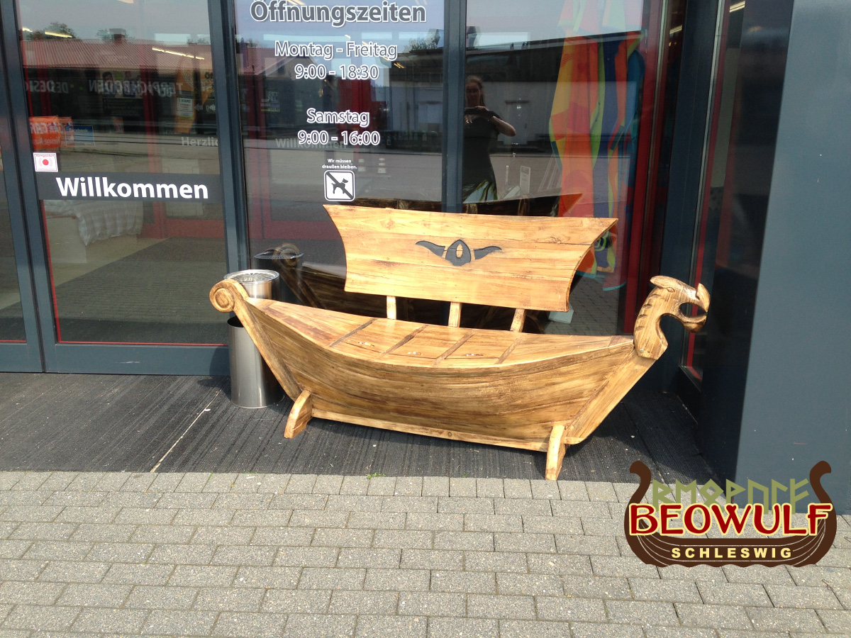 Vor der modernen Glastür eines Geschäftes steht eine Holzbank in Form eines Wikingerschiffes mit Drachenkopf am Bug. Das Segel ist die Lehne.