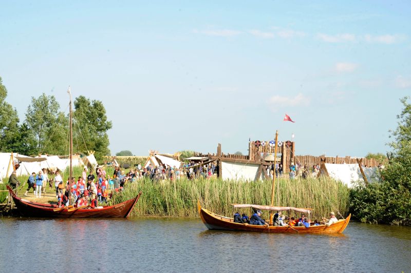 zwei Holzboote mit zusammengelegten Rahsegeln vor einem Anleger im Schilfgürtel, dahinter Menschen zwischen Wikingerzelten und einer Burganlage aus Holzpalisaden